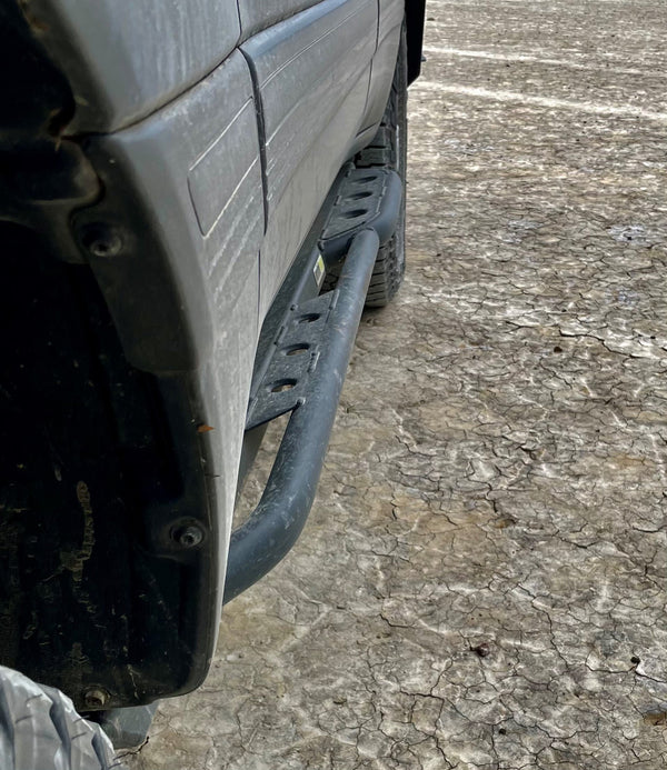 Close-up of black rock slider installed on side of 100 Series Land Cruiser LX470 on cracked dirt ground.