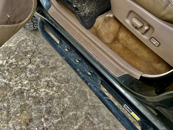 Close-up of black metal rock slider installed on 100 Series Land Cruiser LX470 with tan interior and cracked ground below.
