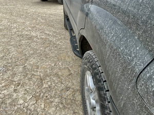 Close-up of black rock slider installed on dirty 100 Series Land Cruiser LX470 side above rugged tire.