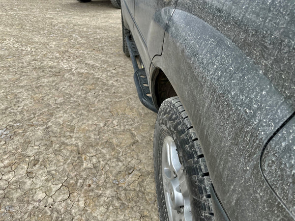 Close-up of black rock slider installed on dirty 100 Series Land Cruiser LX470 side above rugged tire.