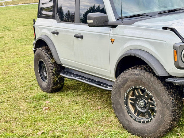Close-up of black rock sliders installed on the driver side of a white 2021 Ford Bronco 4-door on grass.