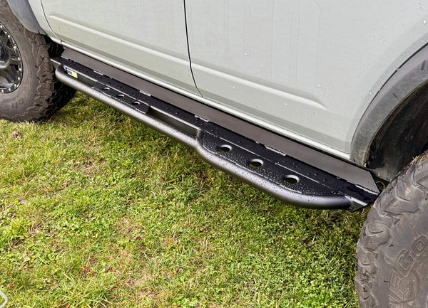 Close-up of black metal rock slider installed on the side of a white 2021 Ford Bronco 4-door on grass.