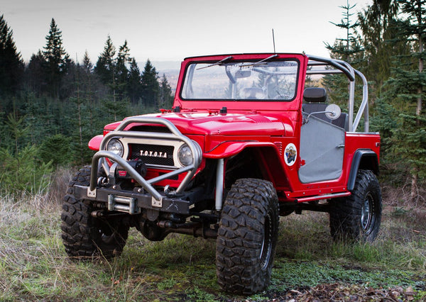 Red Toyota FJ40 Land Cruiser with full-profile metal roll cage and off-road tires parked in forest clearing.