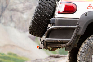Close-up of metal tube rear bumper installed on white off-road vehicle with spare tire and rugged terrain background.