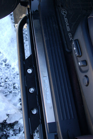 Close-up of black metal rock slider installed on 2010 Lexus GX460 door sill with Lexus floor mat and snow outside.