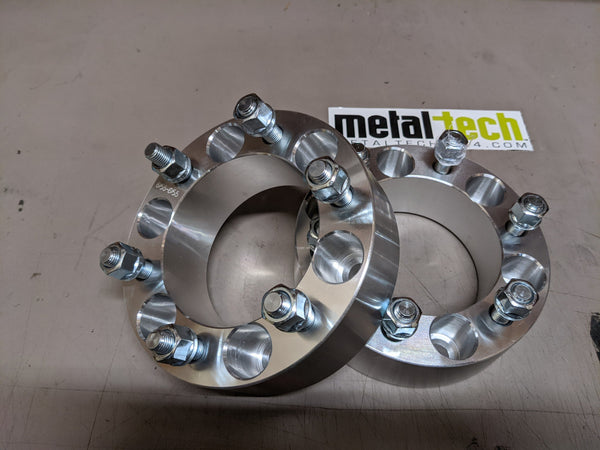 Pair of 6x5.5 bolt pattern, 2-inch thick aluminum wheel spacers with lug studs and nuts on a gray surface.