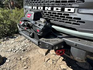 Close-up of Bronco 6G Stronghold winch mount installed on front bumper with Warn winch and rocky terrain background.