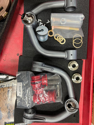 Gray metal upper control arms with hardware and red bushings for 2000-2006 Tundra and 2001-2007 Sequoia, laid out in foam.