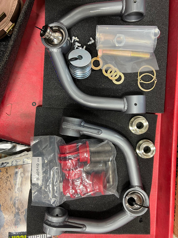 Gray metal upper control arms with hardware and red bushings for 2000-2006 Tundra and 2001-2007 Sequoia, laid out in foam.