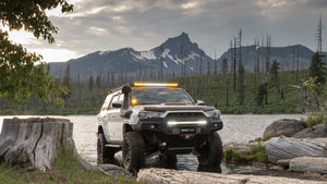 White 5th gen 4Runner with Metal Tech Fortress front bumper and off-road lights, parked by a lake with mountains behind.