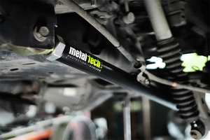 Close-up of black Metal Tech rear lower link installed on vehicle suspension with visible coil spring and shock absorber.