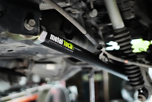 Close-up of black Metal Tech rear lower link installed on vehicle suspension with visible coil spring and shock absorber.